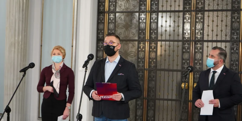 Trzech polityków w maseczkach stoi przy mikrofonach podczas konferencji prasowej w Sejmie, a w tle widoczne są ozdobne drzwi; środkowy mężczyzna trzyma folder z napisem Lewica.
