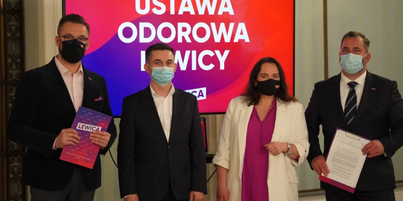 Czterech polityków Lewicy w garniturach i maseczkach stoi na konferencji prasowej, trzymając materiały promocyjne, przed ekranem z napisem „USTAWA ODOROWA LEWICY”.