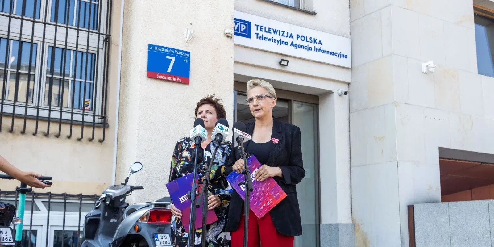 Dwie kobiety stoją przed wejściem do siedziby Telewizji Polskiej i udzielają konferencji prasowej, trzymając materiały Lewicy.