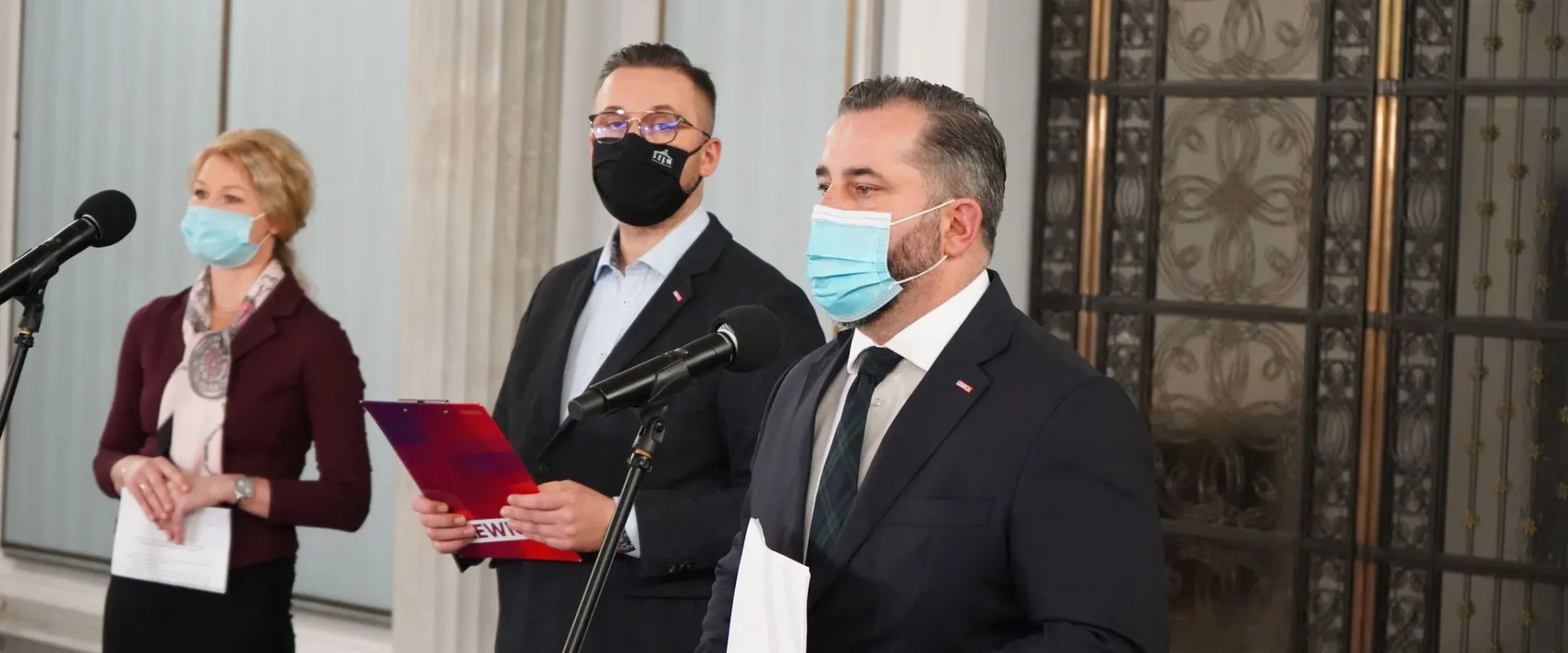 Trzy osoby w maseczkach stoją przy mikrofonach podczas konferencji prasowej w budynku z ozdobnym wejściem, trzymają dokumenty; kontekst to projekt ustawy o 100% wynagrodzenia dla osób w izolacji lub kwarantannie.