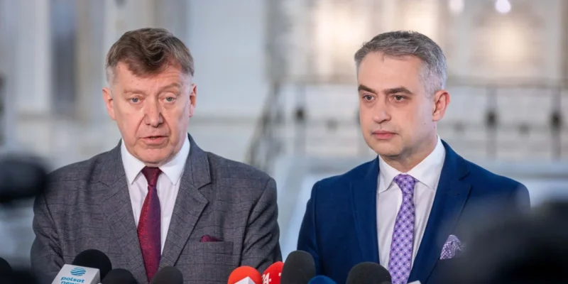 Dwóch mężczyzn w garniturach podczas konferencji prasowej, stojących za licznymi mikrofonami z logo mediów.