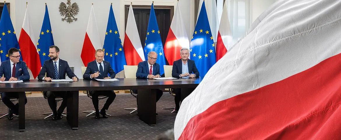 Grupa polityków podpisuje dokumenty przy długim stole, na tle flag Polski i Unii Europejskiej.