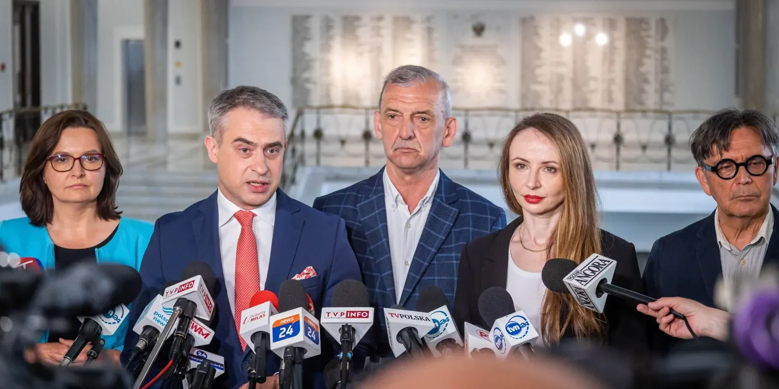 Na zdjęciu widoczna jest grupa osób z mikrofonami, które wydają się dawać oświadczenie w przestrzeni publicznej.