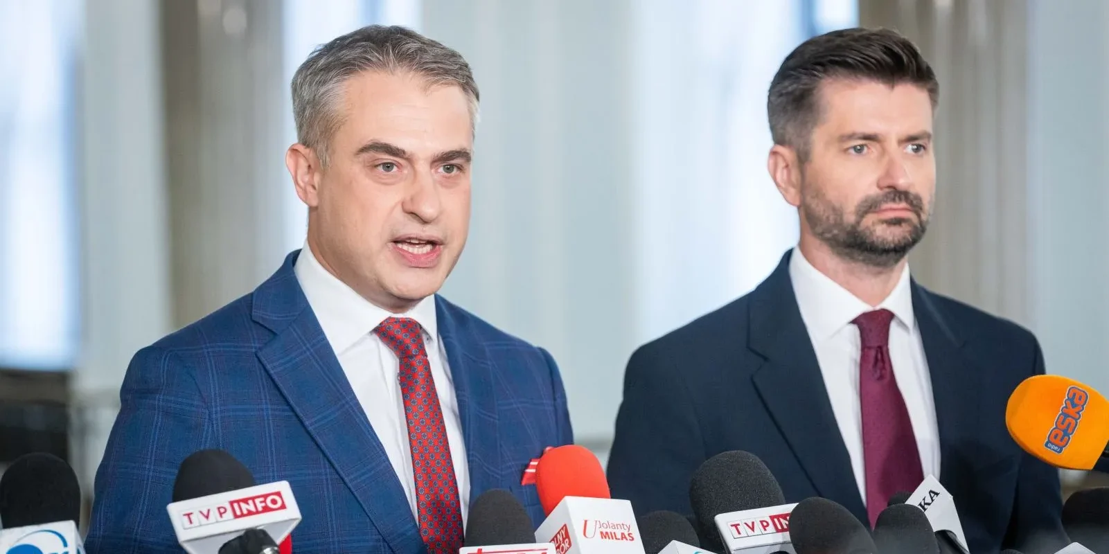 Dwóch mężczyzn w garniturach podczas konferencji prasowej Komisji ds. badania wpływów rosyjskich, stojących obok licznych mikrofonów mediów.