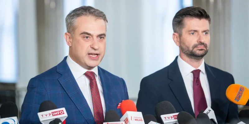 Dwóch mężczyzn w garniturach podczas konferencji prasowej Komisji ds. badania wpływów rosyjskich, stojących obok licznych mikrofonów mediów.