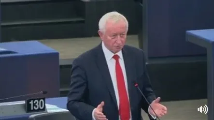 Mężczyzna w ciemnym garniturze i czerwonym krawacie stoi przy mikrofonie, gestykulując podczas wystąpienia na sali obrad.