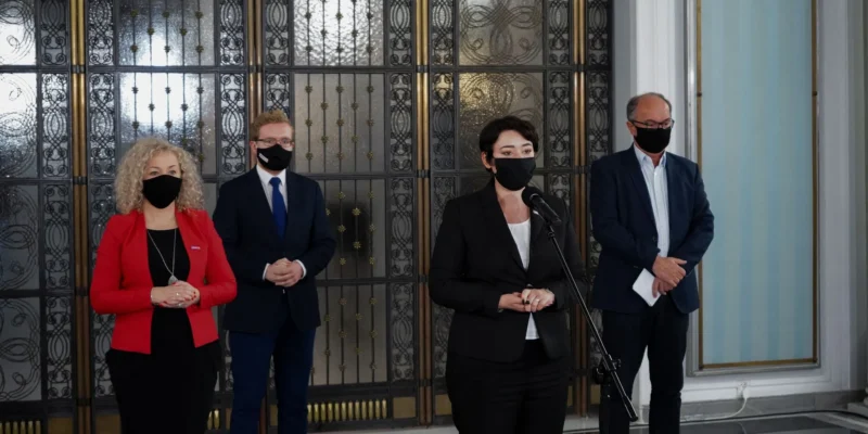 Kobieta w czarnym garniturze przemawia do mikrofonu podczas konferencji prasowej, stojąc między trzema osobami w garniturach, wszyscy w maseczkach, na tle ozdobnej metalowej bramy.