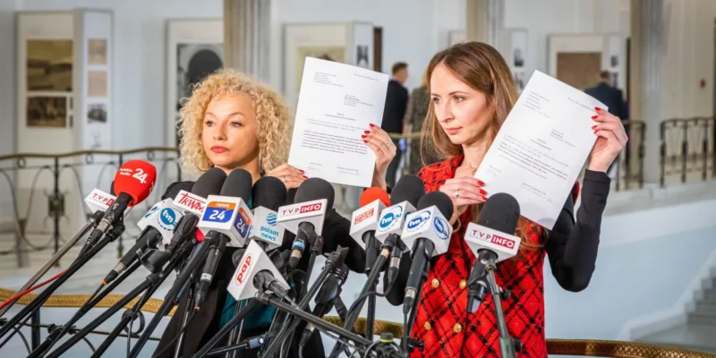 Dwie kobiety trzymające dokumenty stoją przed mikrofonami podczas konferencji prasowej w jasnym wnętrzu.