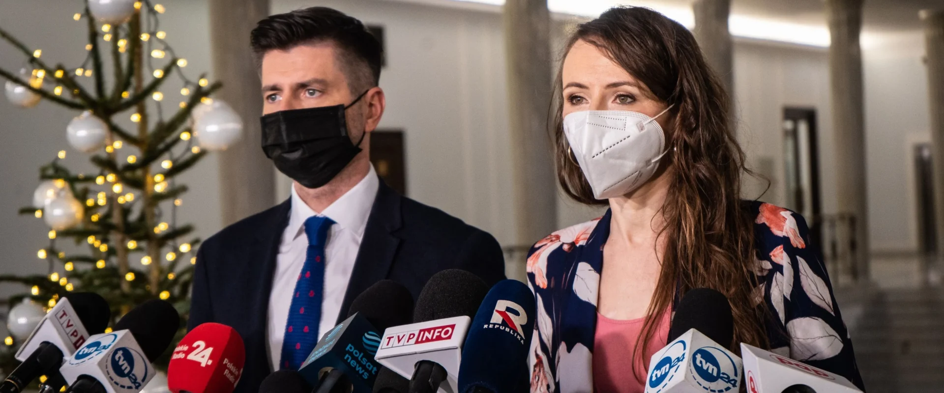 Para polityków w maseczkach stoi na konferencji prasowej otoczona licznymi mikrofonami, a w tle widoczna dekoracja świąteczna z choinką i lampeczkami.