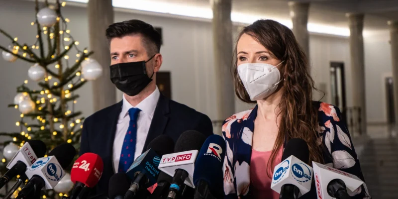 Para polityków w maseczkach stoi na konferencji prasowej otoczona licznymi mikrofonami, a w tle widoczna dekoracja świąteczna z choinką i lampeczkami.