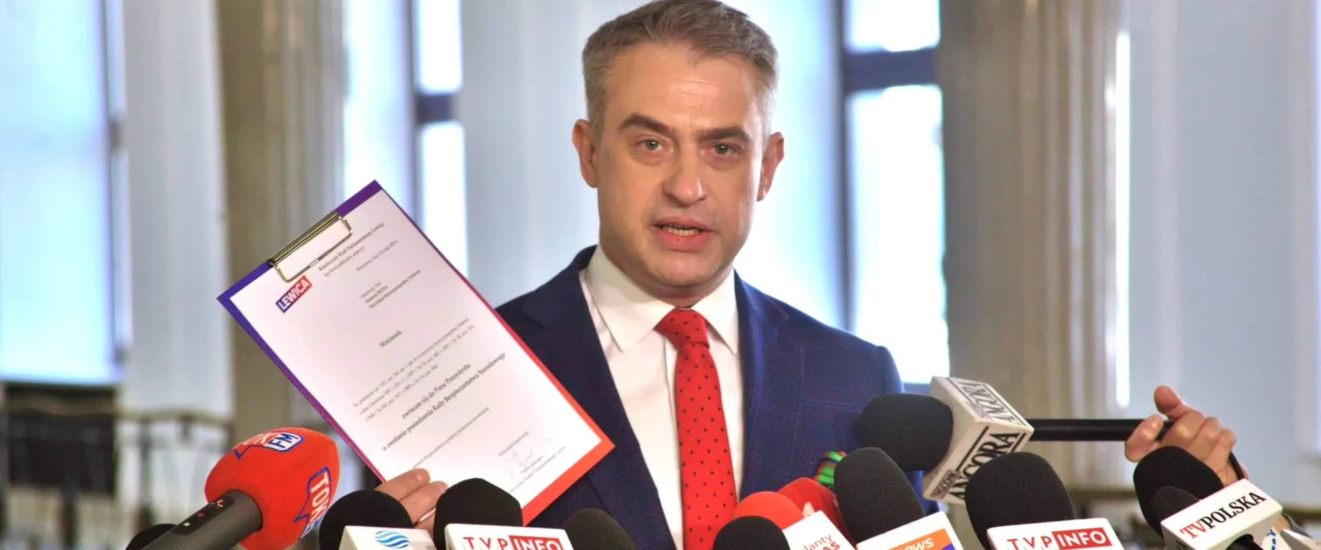 Mężczyzna w garniturze prezentuje dokument podczas konferencji prasowej w otoczeniu mikrofonów różnych stacji telewizyjnych.