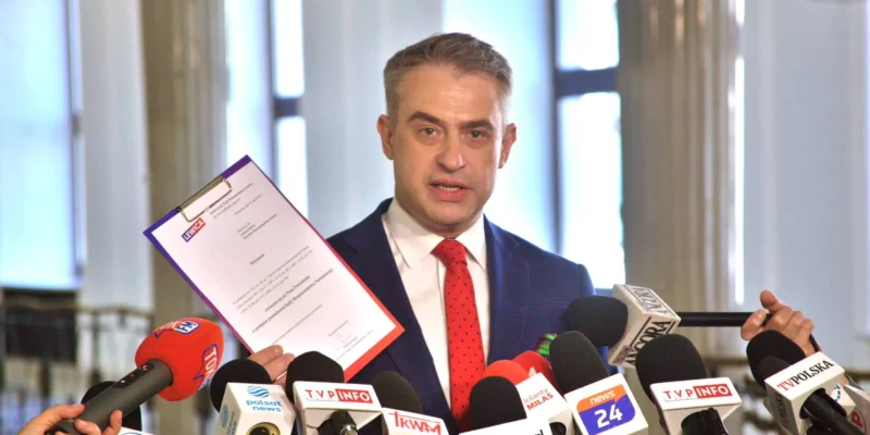 Mężczyzna w garniturze prezentuje dokument podczas konferencji prasowej w otoczeniu mikrofonów różnych stacji telewizyjnych.