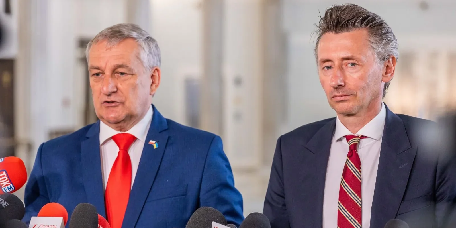 Dwóch mężczyzn w garniturach stoi przed mikrofonami, prowadząc konferencję prasową w jasnej sali.