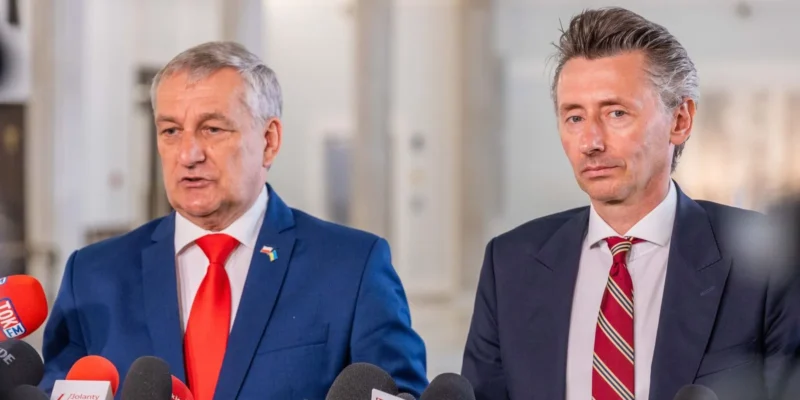 Dwóch mężczyzn w garniturach stoi przed mikrofonami, prowadząc konferencję prasową w jasnej sali.