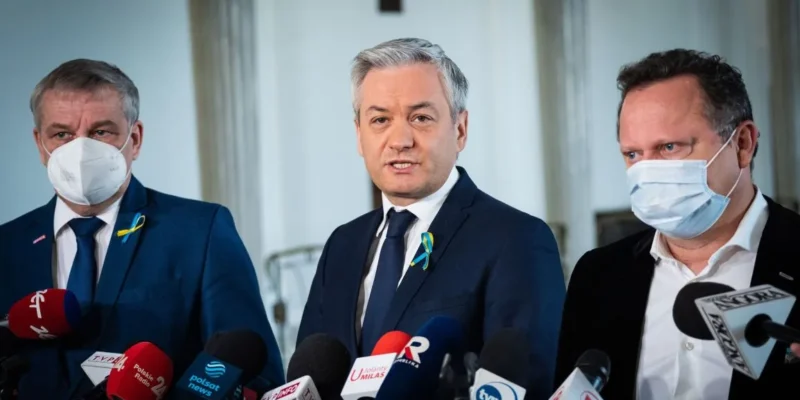 Trzej mężczyźni w garniturach i maseczkach, stojący przed mikrofonami i kamerami, udzielają wywiadu w formalnym otoczeniu.