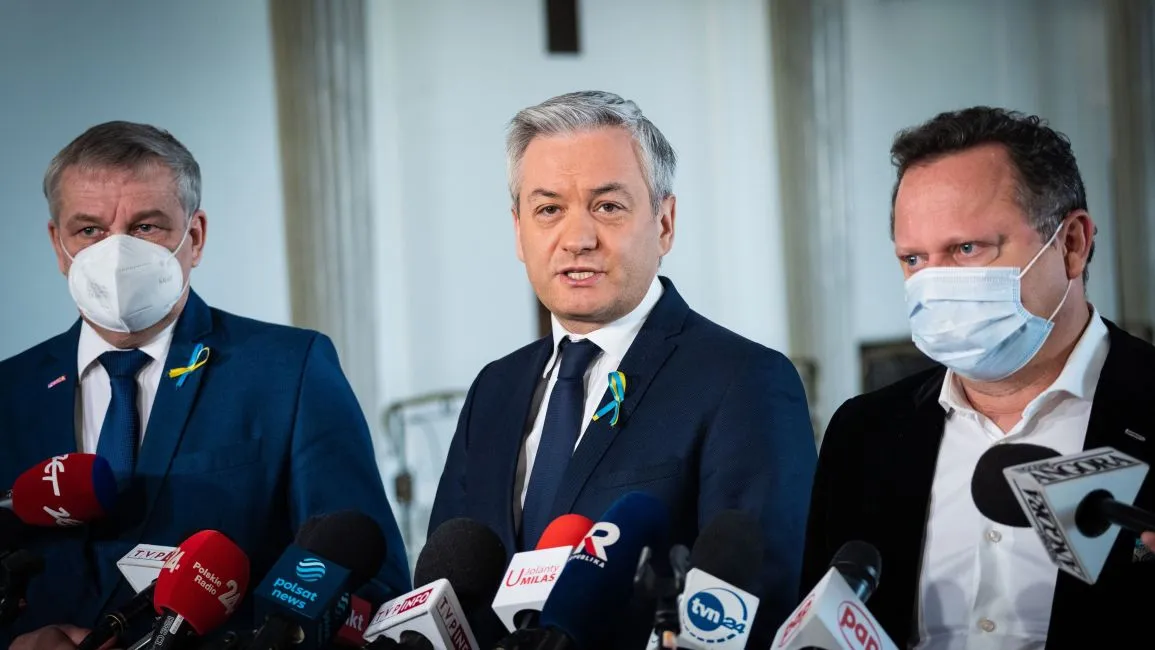 Trzej mężczyźni w garniturach i maseczkach, stojący przed mikrofonami i kamerami, udzielają wywiadu w formalnym otoczeniu.