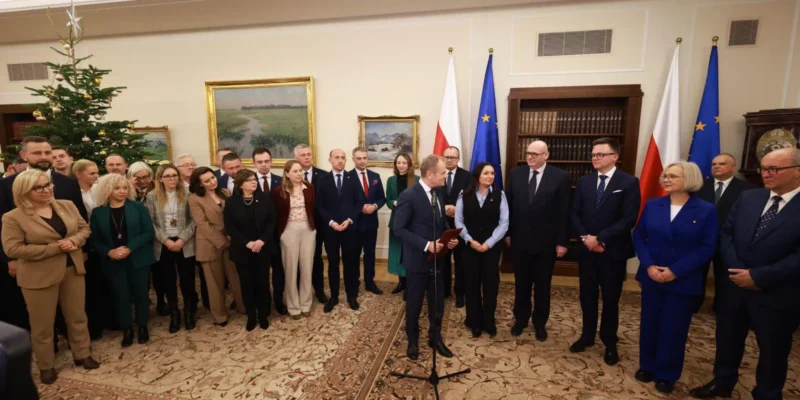 Grupa polityków w eleganckich strojach stoi w reprezentacyjnej sali z choinką i obrazami na ścianach, a mężczyzna przy mikrofonie przemawia, na tle flag Polski i Unii Europejskiej.