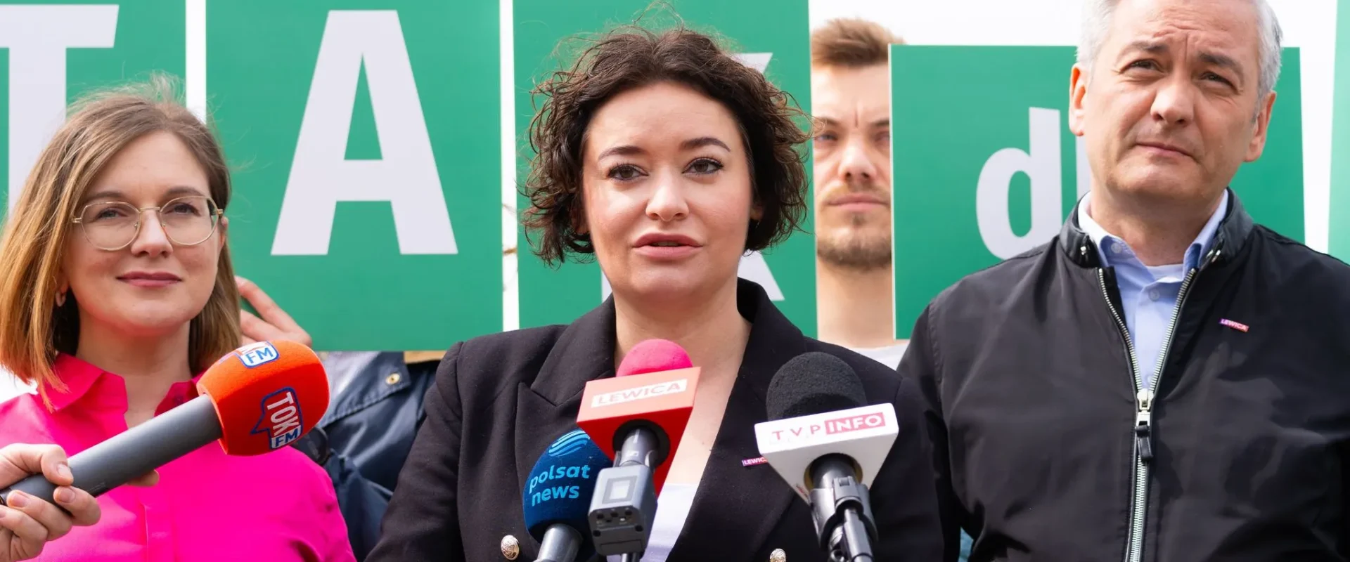 Grupa osób na konferencji prasowej stoi przed zielonymi tablicami z literami TAK w tle, a osoba przemawia do mikrofonów.
