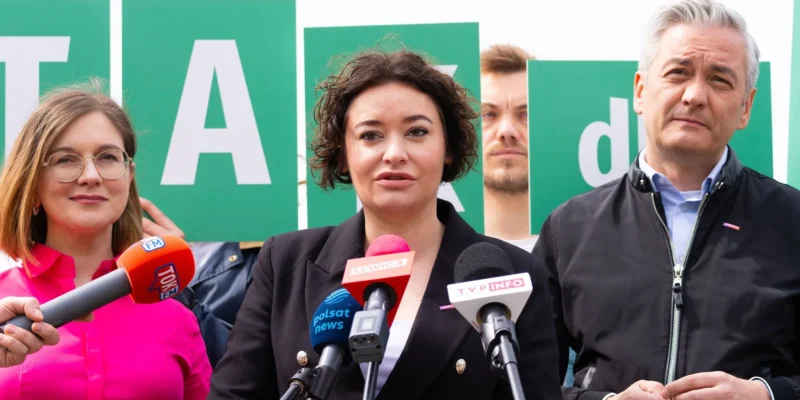 Grupa osób na konferencji prasowej stoi przed zielonymi tablicami z literami TAK w tle, a osoba przemawia do mikrofonów.