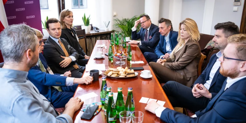 Grupa polityków w garniturach siedzi przy długim stole w sali konferencyjnej, rozmawiając przy napojach i przekąskach, a w tle widać baner Lewicy symbolizujący wsparcie dla wolnej Białorusi.