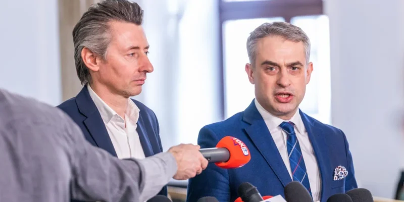 Dwaj mężczyźni w garniturach rozmawiają z dziennikarzami podczas konferencji prasowej, a w tle znajdują się mikrofony.