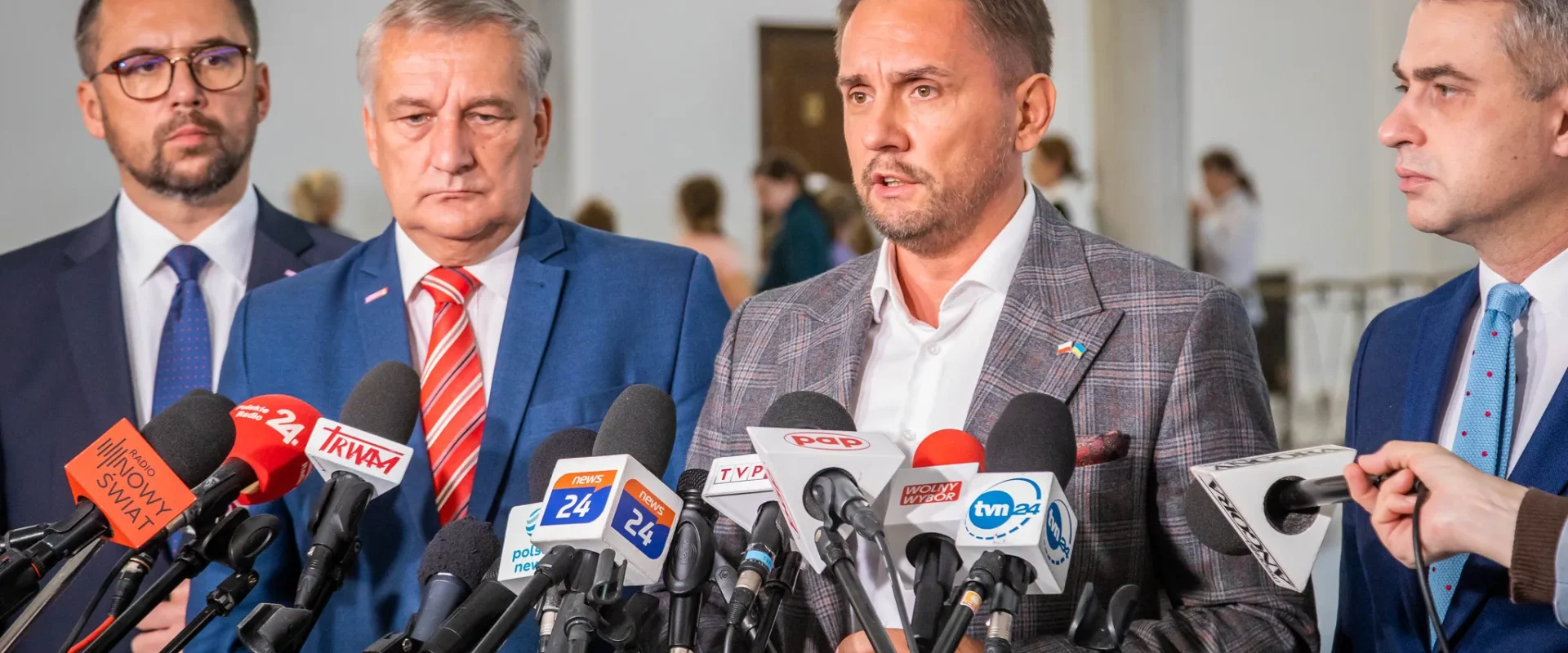 Na zdjęciu czterech mężczyzn w garniturach udziela wywiadu przed grupą mikrofonów w pomieszczeniu publicznym.