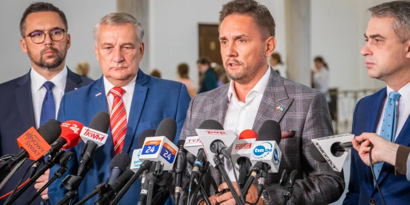 Na zdjęciu czterech mężczyzn w garniturach udziela wywiadu przed grupą mikrofonów w pomieszczeniu publicznym.