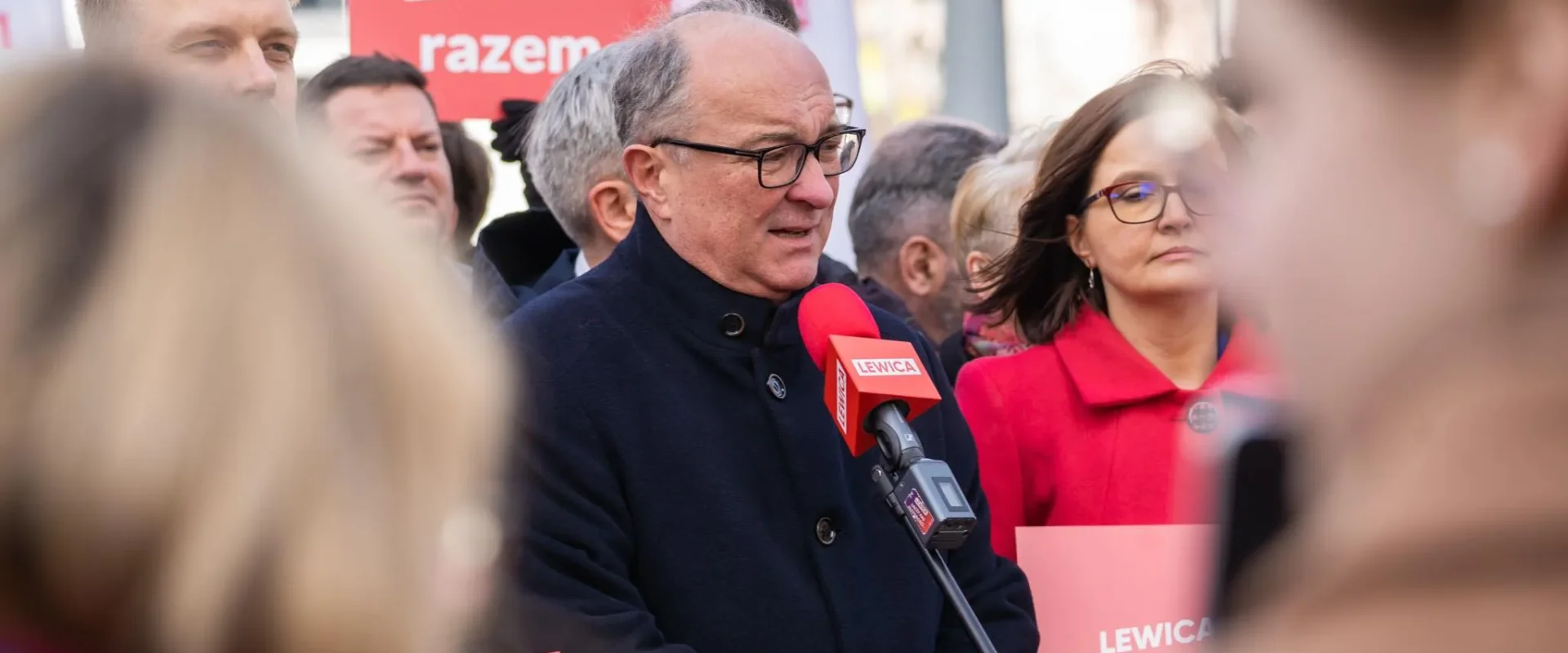 Na zdjęciu widoczna jest grupa osób biorących udział w wydarzeniu politycznym, z mikrofonem w dłoni wypowiada się mężczyzna w ciemnym płaszczu.