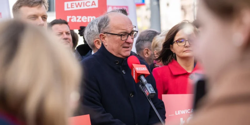 Na zdjęciu widoczna jest grupa osób biorących udział w wydarzeniu politycznym, z mikrofonem w dłoni wypowiada się mężczyzna w ciemnym płaszczu.