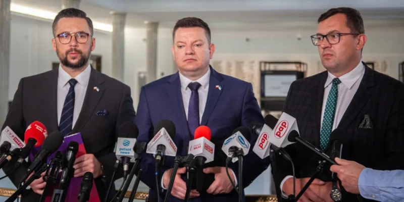 Trzech mężczyzn w garniturach stoi na konferencji prasowej otoczonych mikrofonami z logotypami mediów.