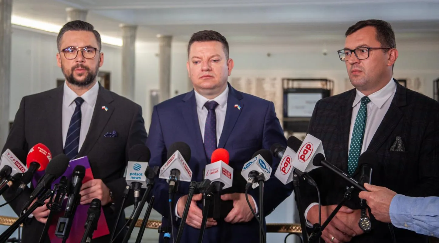 Trzech mężczyzn w garniturach stoi na konferencji prasowej otoczonych mikrofonami z logotypami mediów.