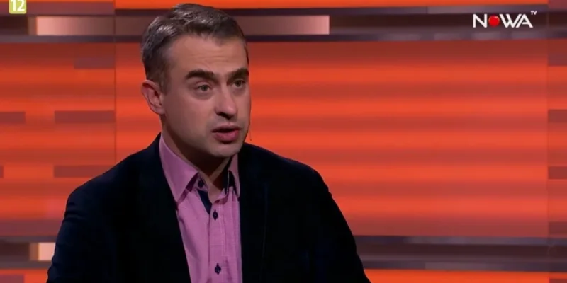 Mężczyzna w ciemnej marynarce i różowej koszuli mówi w studiu telewizyjnym na pomarańczowym tle z widocznym logo stacji.