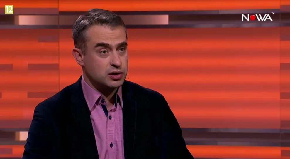 Mężczyzna w ciemnej marynarce i różowej koszuli mówi w studiu telewizyjnym na pomarańczowym tle z widocznym logo stacji.