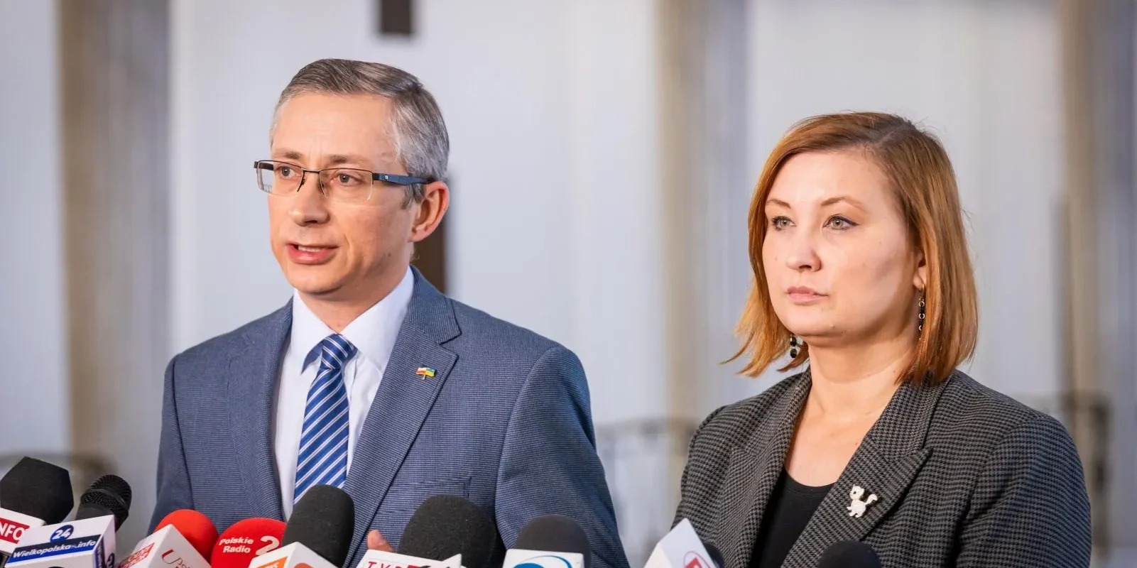 Dwóch urzędników (mężczyzna w garniturze i kobieta w marynarce) stoi przy licznych mikrofonach podczas konferencji prasowej dotyczącej regionalnego okrągłego stołu o problemach w psychiatrii dziecięcej w Białymstoku, Elblągu, Gdyni, Łodzi, Opolu, Radomiu i we Wrocławiu.