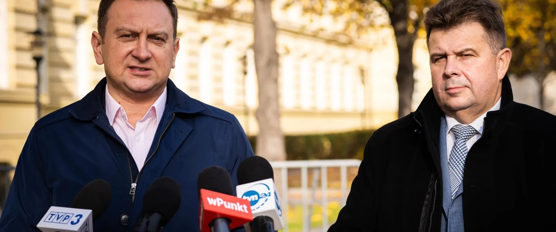 Dwaj mężczyźni w garniturach stoją przy mikrofonach, na tle jesiennego krajobrazu.