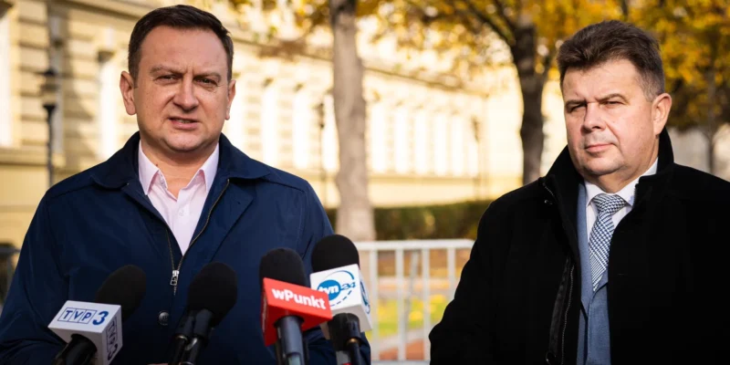 Dwaj mężczyźni w garniturach stoją przy mikrofonach, na tle jesiennego krajobrazu.