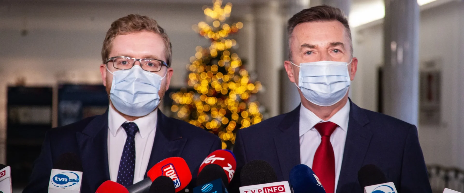 Dwóch mężczyzn w maseczkach rozmawia z dziennikarzami, w tle widoczne ozdobione drzewko świąteczne.