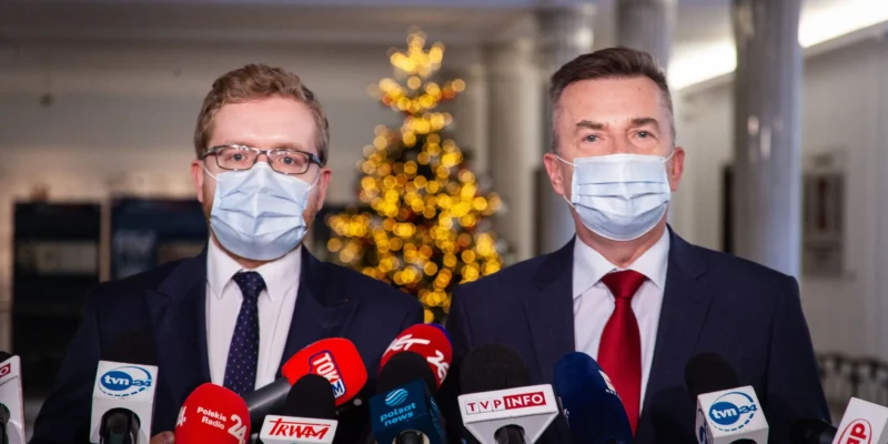 Dwóch mężczyzn w maseczkach rozmawia z dziennikarzami, w tle widoczne ozdobione drzewko świąteczne.