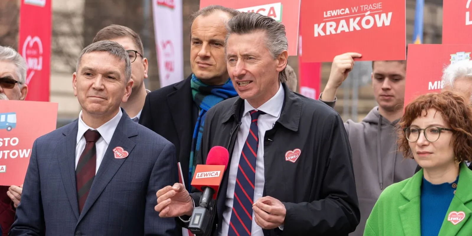 Politycy Lewicy podczas konferencji prasowej, stoją przy mikrofonie z napisem LEWICA, wokół czerwone plakaty i banery Lewicy.