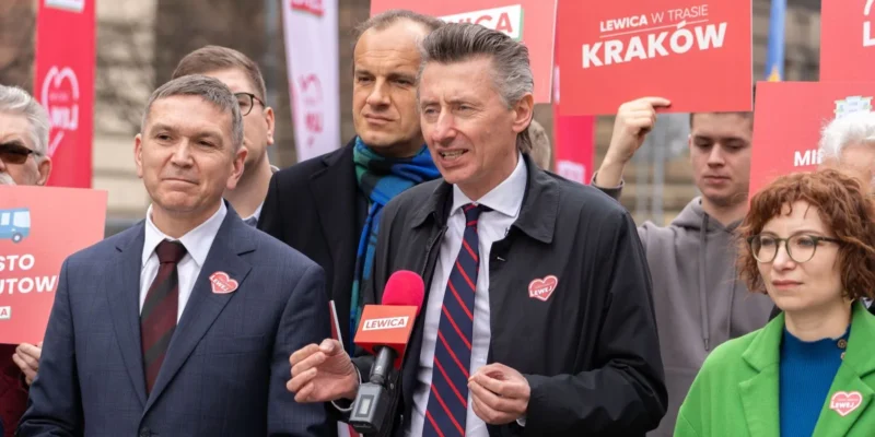 Politycy Lewicy podczas konferencji prasowej, stoją przy mikrofonie z napisem LEWICA, wokół czerwone plakaty i banery Lewicy.