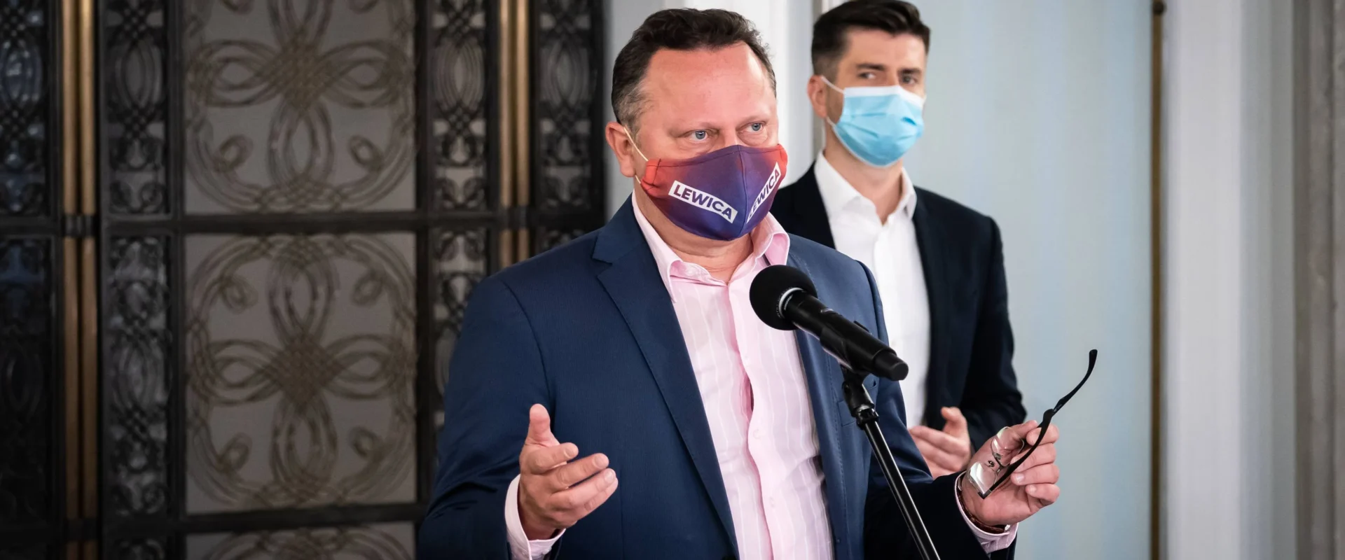 Mężczyzna w granatowym garniturze i różowej koszuli przemawia do mikrofonu, nosząc maseczkę z napisem Lewica, podczas gdy za nim stoi drugi mężczyzna w niebieskiej maseczce.