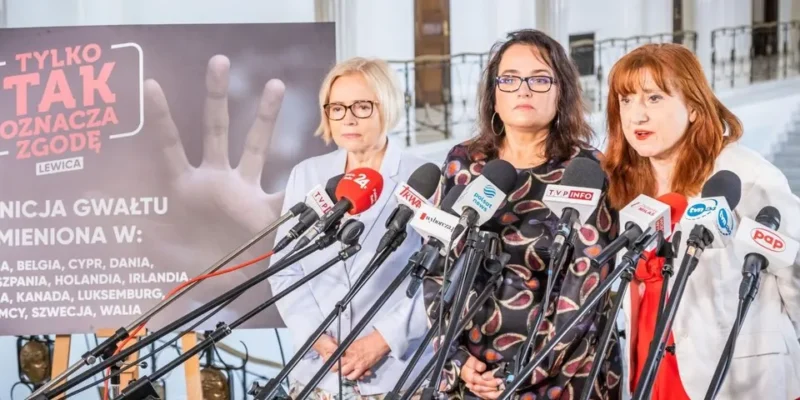 Trzy kobiety stoją przed mikrofonami w sali konferencyjnej, prezentując temat związany z definicją zgody i przemocy w kontekście ustawodawstwa.