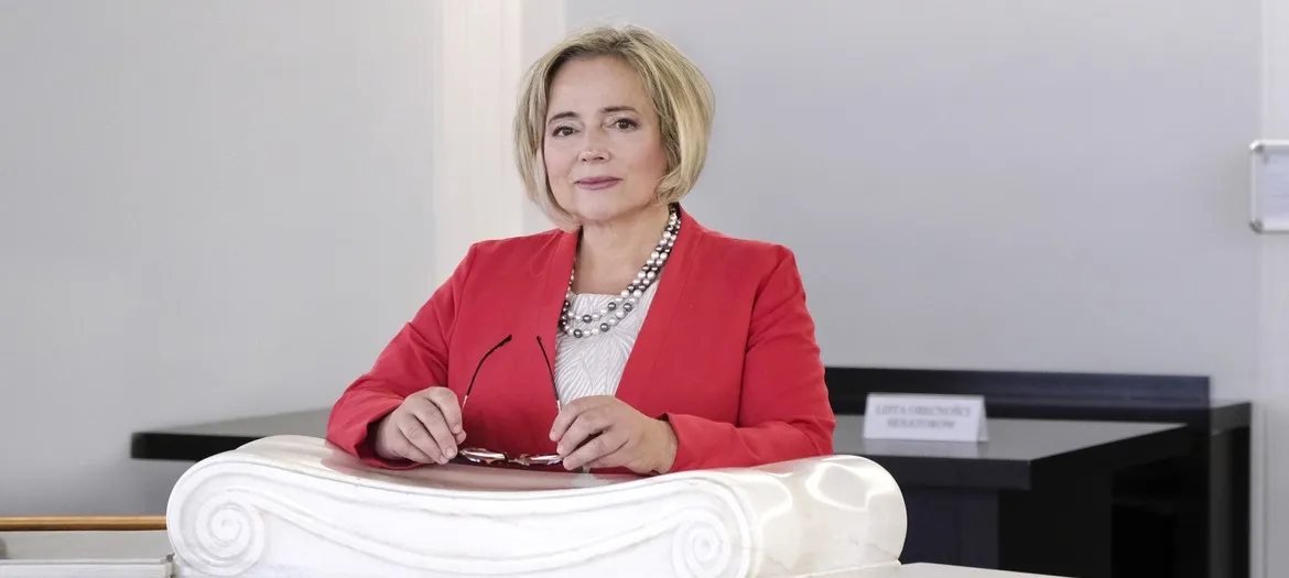 Kobieta w czerwonej marynarce siedzi za biurkiem i trzyma okulary, na tle neutralnego biurowego wnętrza.