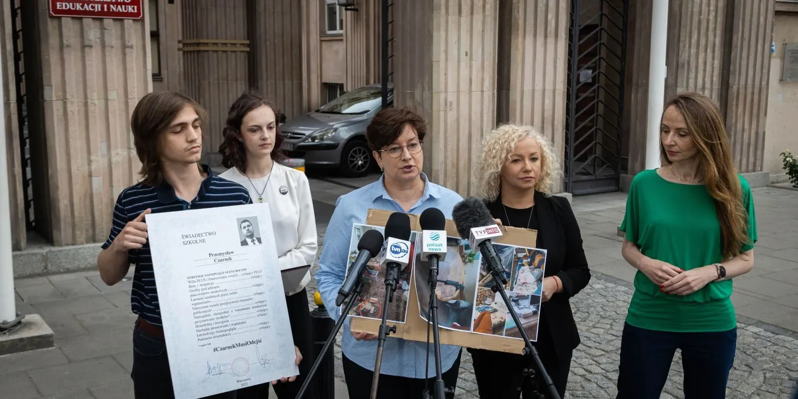 Grupa osób podczas konferencji prasowej przed siedzibą Ministerstwa Edukacji i Nauki, z mikrofonami skierowanymi ku mówiącemu, omawia decyzję o nieprzyznaniu promocji do następnej klasy.