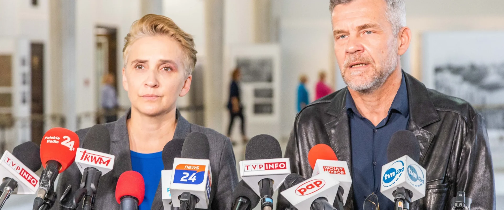 Dwóch polityków stoi przed zebranym tłumem dziennikarzy, otoczeni mikrofonami, w tle można dostrzec wystawę.