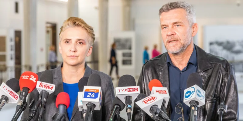 Dwóch polityków stoi przed zebranym tłumem dziennikarzy, otoczeni mikrofonami, w tle można dostrzec wystawę.