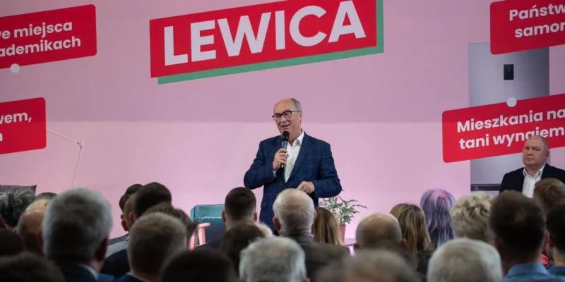 Mężczyzna w granatowym garniturze przemawia do zgromadzonej publiczności na scenie, na różowym tle z dużym napisem LEWICA.