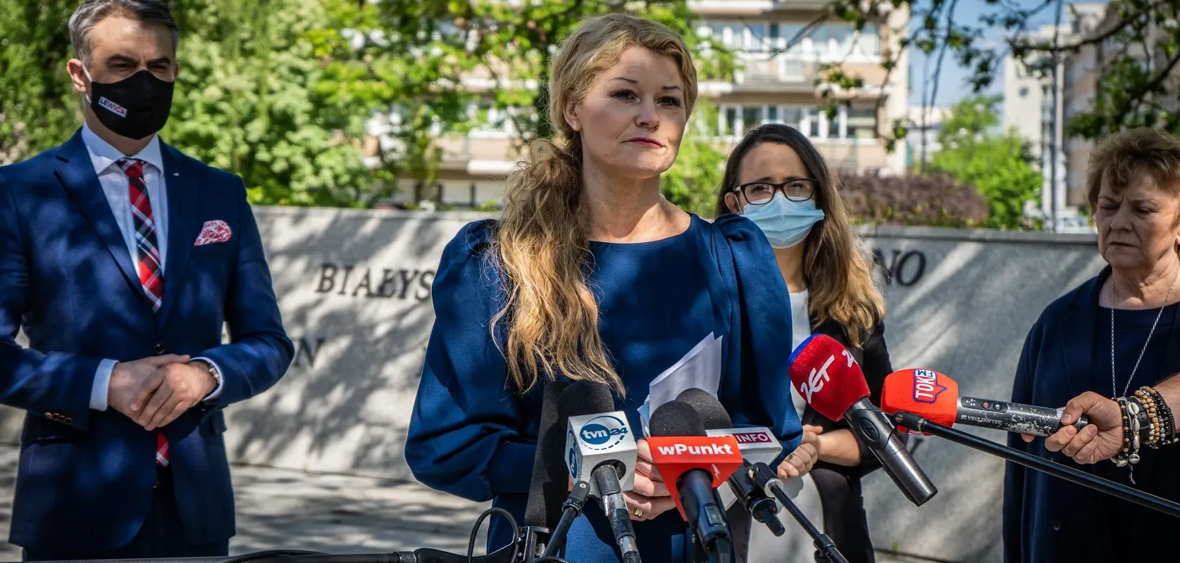 Kobieta w niebieskim kostiumie przemawia na konferencji prasowej, stojąc za licznymi mikrofonami i w otoczeniu dziennikarzy.