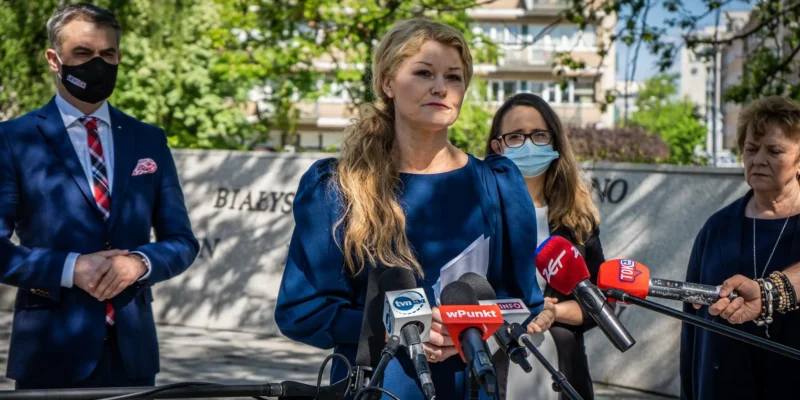 Kobieta w niebieskim kostiumie przemawia na konferencji prasowej, stojąc za licznymi mikrofonami i w otoczeniu dziennikarzy.