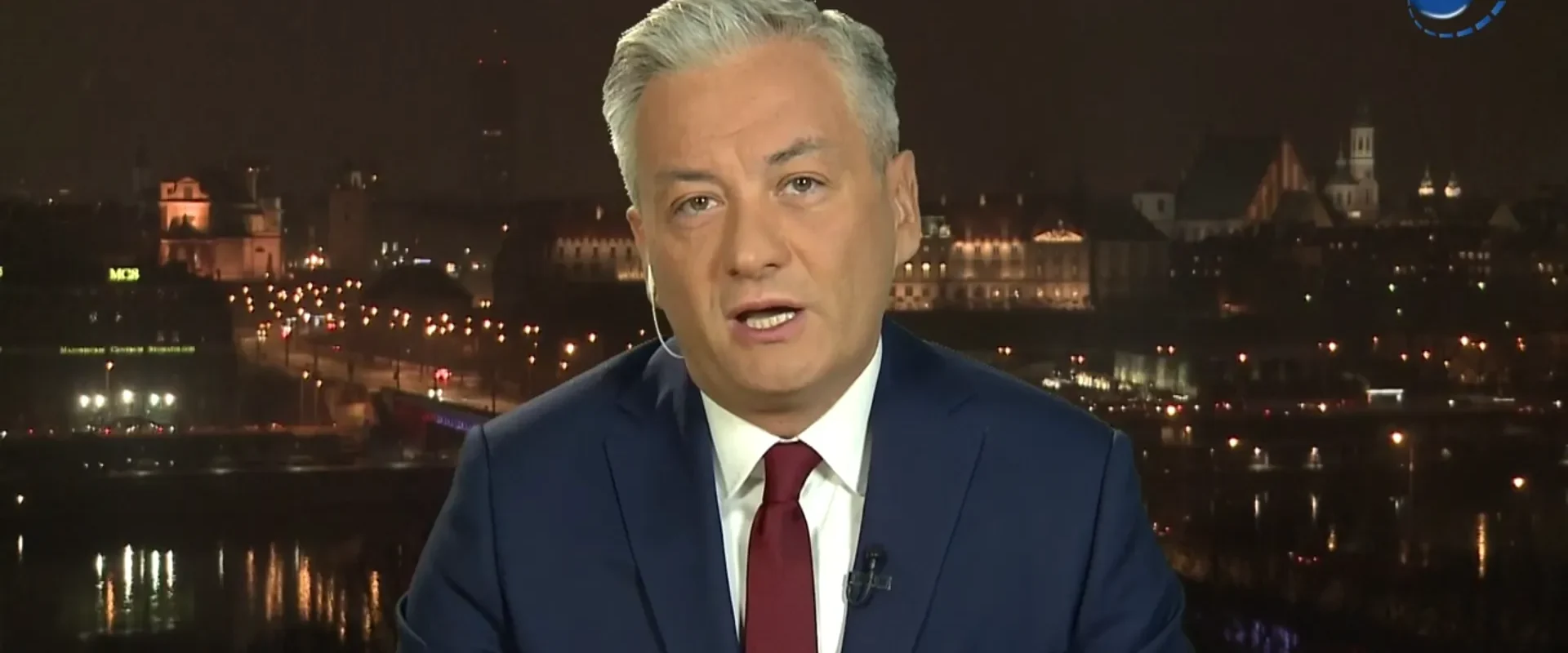 Dziennikarz w granatowym garniturze i czerwonym krawacie przemawia do kamery na tle nocnego miasta; w prawym górnym rogu widoczny jest logotyp TVN24.